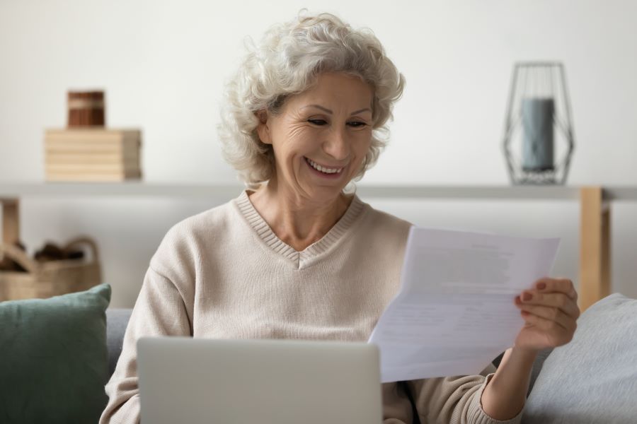 Happy senior woman with laptop reading paper letter at home